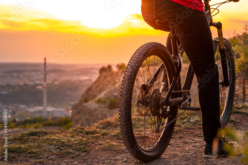 Back shot of mountain biker at sunset sitting on a modern off-road bike on the hill behind the city.