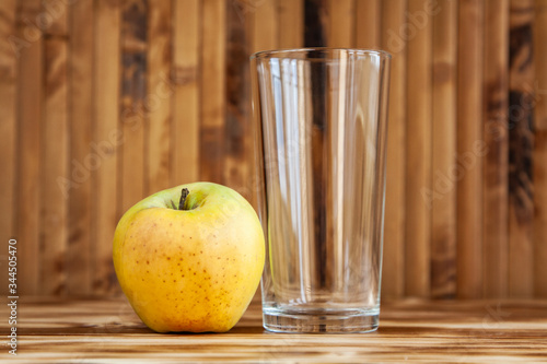 A glass cup stands on a natural wood countertop against a bamboo wall, next to it is a yellow apple