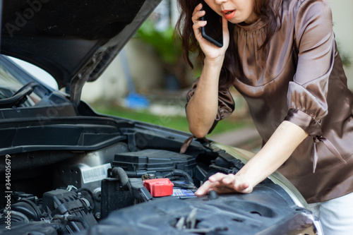 asia angry woman looking at broken down car engine and holding mobile phone calling for help