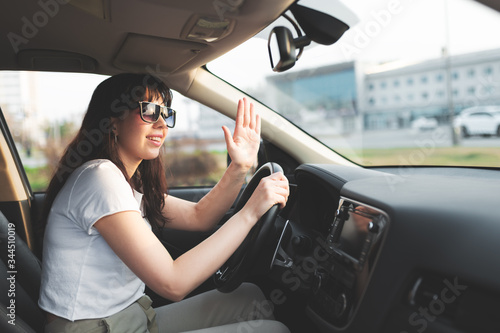 Pretty brunette in white tshirt and sunglasses sitting on the drivers sitting and welcome someone throught the windshield.  Woman in car waving hand to a pedestrian.