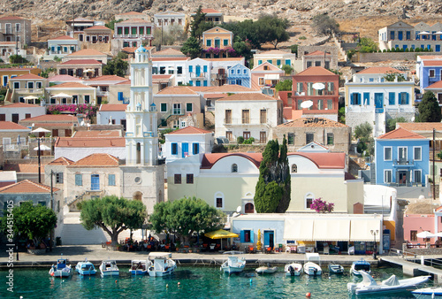 The Greek holiday island of Halki. A view of the bay at the port of Emborio. Pleasure boats are moored at the quay. Classically styled houses in pastel colors rise up the hillside.