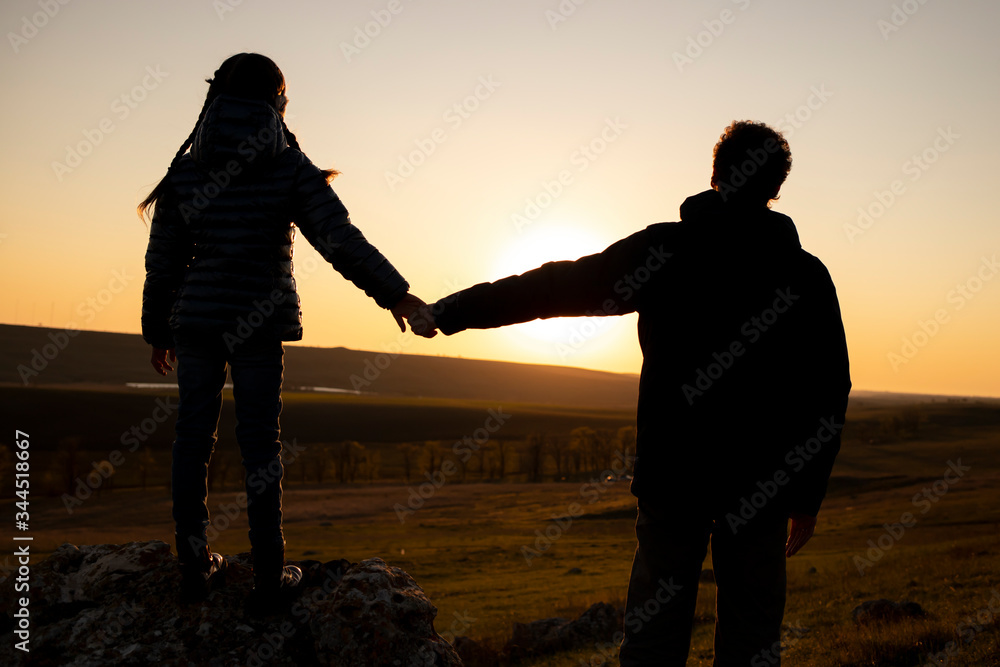 A silhouette of a father and daughter who hold each other's hands against a sunset sky.