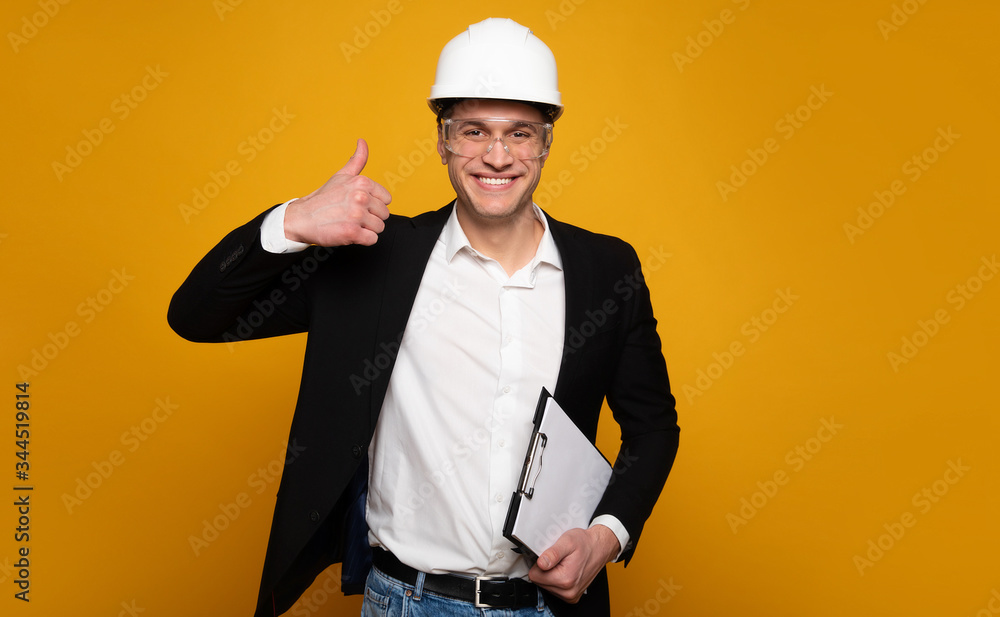 Everything is great. Happy attractive architect in smart casual outfit and white helmet is looking in the camera and showing thumbs up, while holding his notes. Everything is great. Happy attractive architect in smart casual outfit and white helmet is looking in the camera and showing thumbs up, while holding his notes.