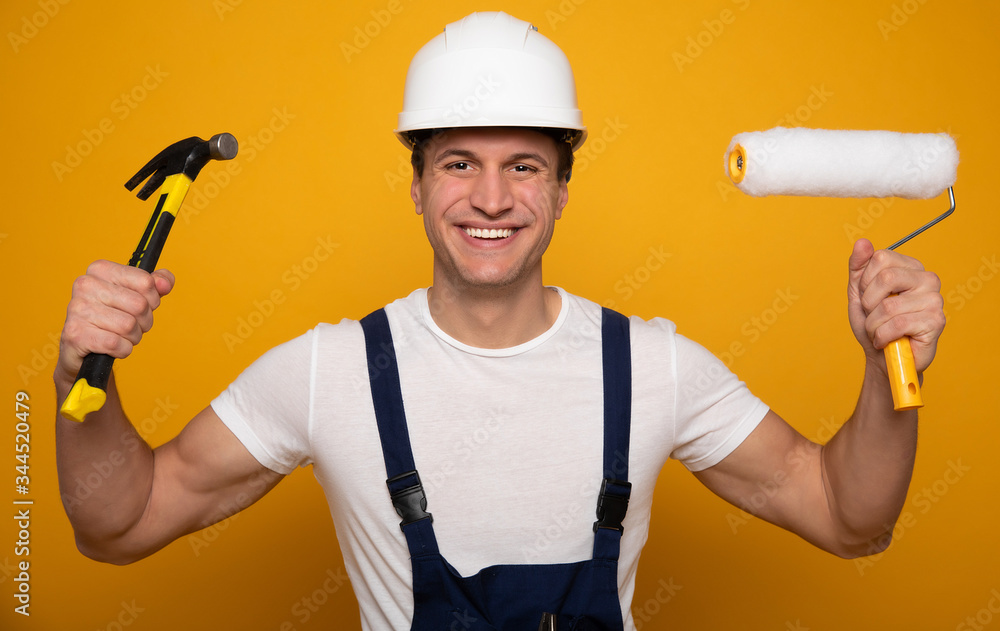 Construction worker. Close-up photo of attractive handyman, who is holding a hammer in his right hand and a painting roller in his left hand, looking in the camera happily.
