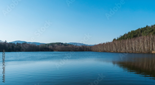 Blue calm water of forest lake, fish pond Kunraticky rybnik with birch and spruce trees growing along the shore and clear blue sky. Nature background. Early spring landscape.