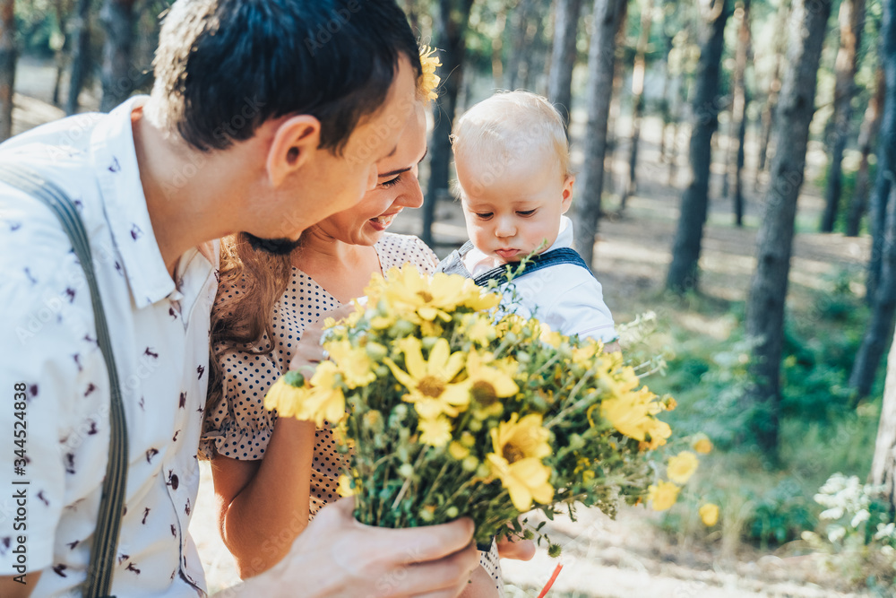 Happy young family spending time outdoor on a summer day. Mom holding yellow daisies in hands. The son carefully looking to flowers. Parents are smiling at their child and enjoying a warm evening