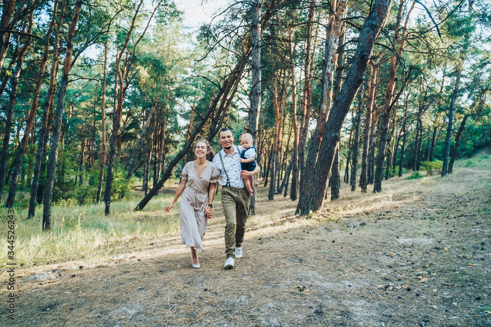 Happy young family having fun running through the evening summer forest. Dad holding in his hands a one-year-old child boy. Parents happily bouncing and having a good time out of town