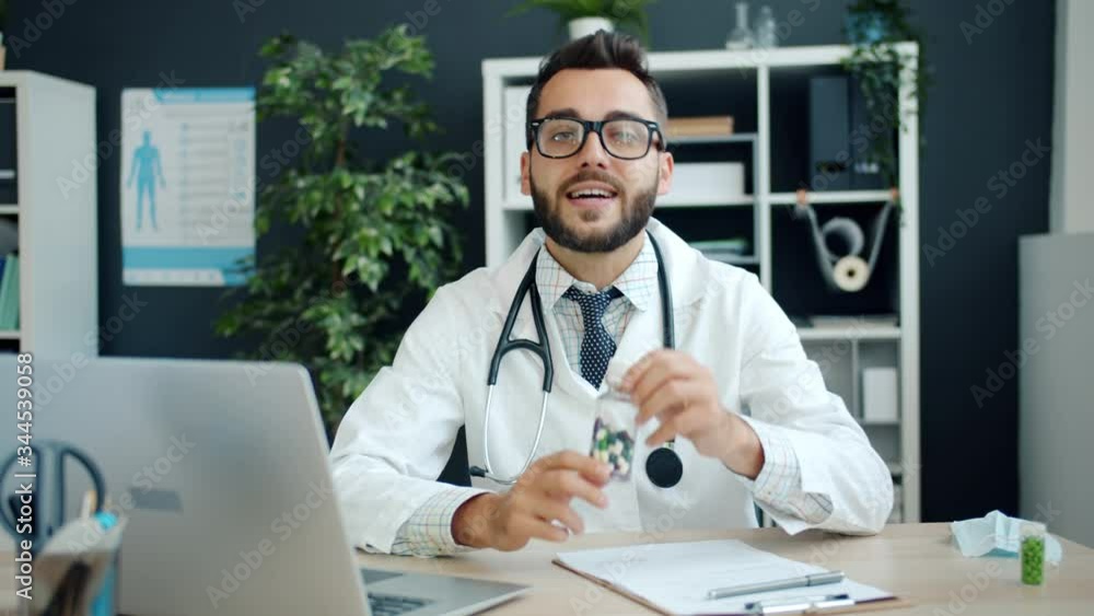 Portrait of handsome young man doctor talking and gesturing showing pills using laptop webcam online call sitting at work table in hospital. Medication, medical specialists and clinic concept. Portrait of handsome young man doctor talking and gesturing showing pills using laptop webcam online call sitting at work table in hospital. Medication, medical specialists and clinic concept.