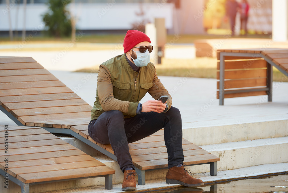 A man in a medical face mask to avoid the spread coronavirus holding a smartphone in the cozy street. A guy turned sideways sitting in a wooden deck chair wears sunglasses and face mask against COVID A man in a medical face mask to avoid the spread coronavirus holding a smartphone in the cozy street. A guy turned sideways sitting in a wooden deck chair wears sunglasses and face mask against COVID
