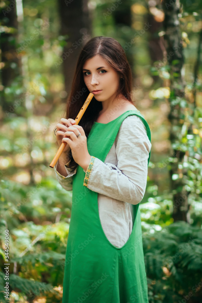 Woman in a green medieval dress plays the wooden flute while standing in a grove against the background of pines. Girl in a gloomy forest plays music on a handmade pipe. Young witch in a gloomy forest Woman in a green medieval dress plays the wooden flute while standing in a grove against the background of pines. Girl in a gloomy forest plays music on a handmade pipe. Young witch in a gloomy forest