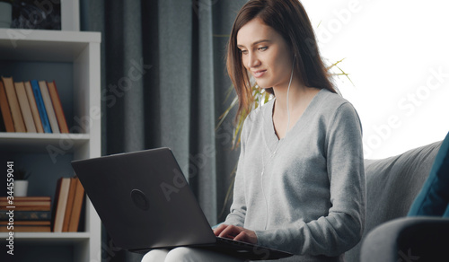 Half-length portrait of young pleased female sitting on sofa with laptop on her lap typing keyborad photo