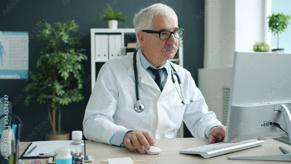 Serious serior man doctor is using computer typing sitting at desk in hospital office working alone. Modern technology, profession and medicine concept.
