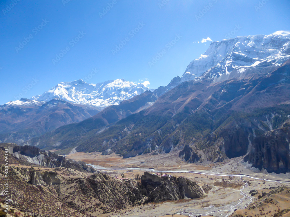 A panoramic view on Manang valley from Praken Gompa, Nepal. High Himalayan ranges around. There is a small torrent in the valley. Snow capped peaks of Annapurna Chain. Harsh landscape.