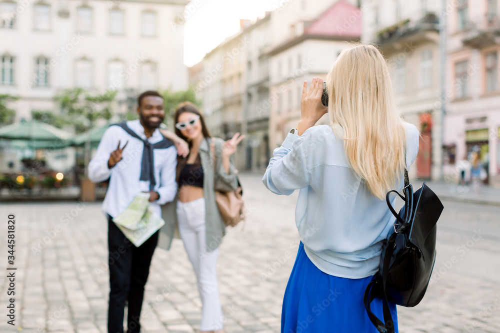 Fototapeta premium Summer holidays, technology and city travel concept. Back view of pretty Caucasian blond girl taking photo of two multiethnic friends, dark skinned man and Caucasian woman in the city