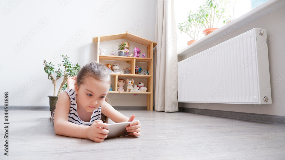 Distance learning. Little girl lying on a wooden floor with a phone, watching a cartoon or making a video call on a computer Distance learning. Little girl lying on a wooden floor with a phone, watching a cartoon or making a video call on a computer