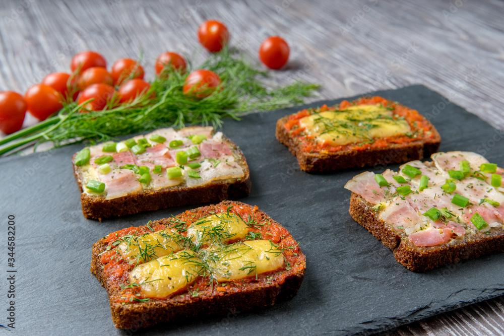Hot sandwiches of rye bread slices with bacon, cheese, green onions and dill on stone board. Concept of fast food and nutritious snacks