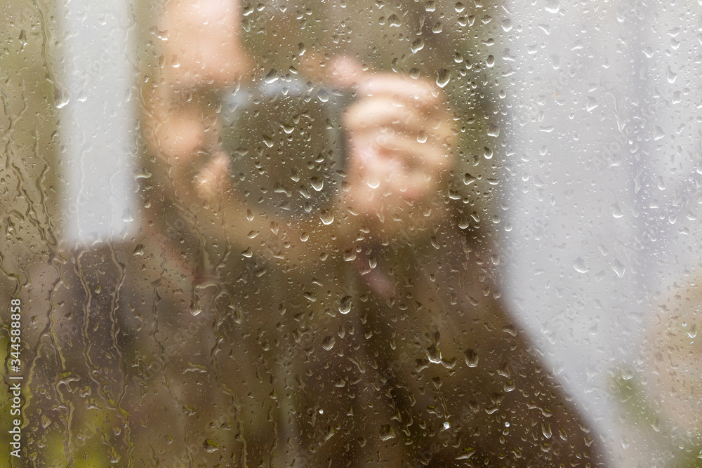View through a glass with raindrops on a man with a camera. View of a person is not in focus, blurred.  Focus on raindrops