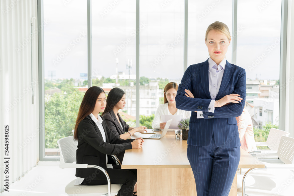 Portrait of Business woman leader self-confident standing in a busy modern workplace while Group of young business people working and communicating sitting at office desk together. union feminists Portrait of Business woman leader self-confident standing in a busy modern workplace while Group of young business people working and communicating sitting at office desk together. union feminists