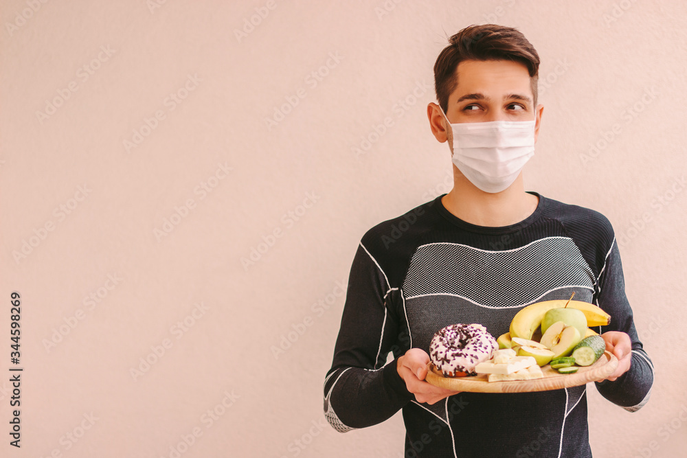 Hungry fitness man in medical face mask hold wooden board with fruits, vegetables, sweet donut, chocolate and look at copy space. Sports man in protective mask dieting at home quarantine. COVID-19 Hungry fitness man in medical face mask hold wooden board with fruits, vegetables, sweet donut, chocolate and look at copy space. Sports man in protective mask dieting at home quarantine. COVID-19