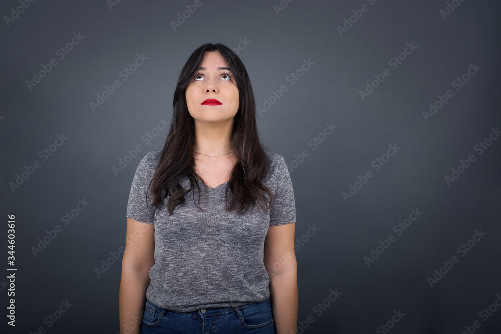 portrait of mysterious charming female with straight hair looking up with enigmatic smile. Beautiful smiling girl looking up standing against gray wall. portrait of mysterious charming female with straight hair looking up with enigmatic smile. Beautiful smiling girl looking up standing against gray wall.