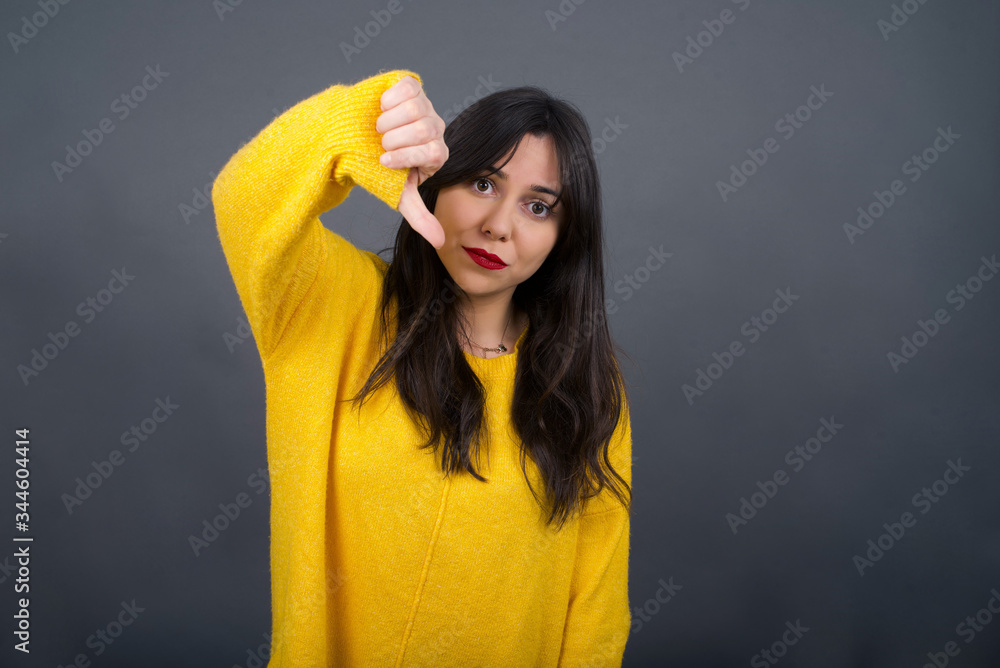 Discontent European woman shows disapproval sign, keeps thumb down, expresses dislike, frowns face in discontent, dressed in white shirt, isolated over gray background. Body language concept. Discontent European woman shows disapproval sign, keeps thumb down, expresses dislike, frowns face in discontent, dressed in white shirt, isolated over gray background. Body language concept.