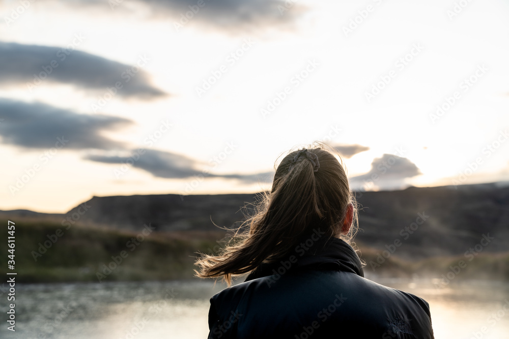 Young girl with brown hair in a ponytail gazing over a lake and a mountain into the sunset. Her hair blow in the wind and the sunlight contours the ponytail. 