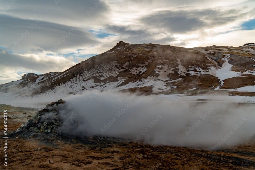 A pile of rocks over a geothermal spring emitting thick warm steam. A muddy partly snow covered mountain in the background. A pile of rocks over a geothermal spring emitting thick warm steam. A muddy partly snow covered mountain in the background.