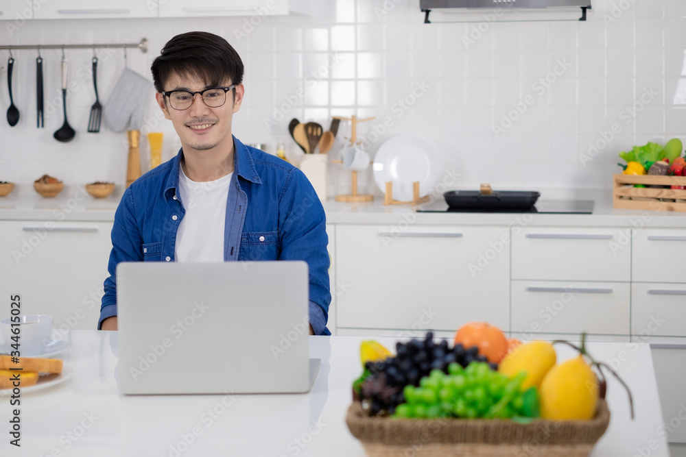 Asian freelance young man working from home with breakfast bread and coffee using laptop in white kitchen Asian freelance young man working from home with breakfast bread and coffee using laptop in white kitchen