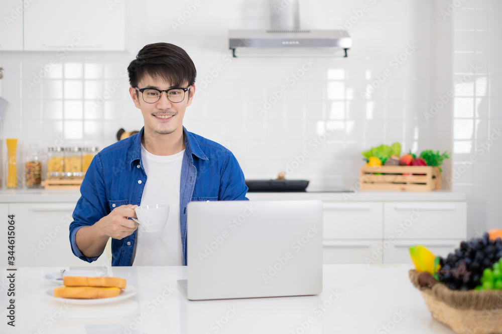 Asian freelance young man working from home with breakfast bread and coffee using laptop in white kitchen Asian freelance young man working from home with breakfast bread and coffee using laptop in white kitchen