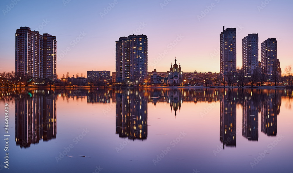 Night city on the lake. Reflected buildings. Lilac sunset. Church between the houses. High-rise buildings. Night lights.