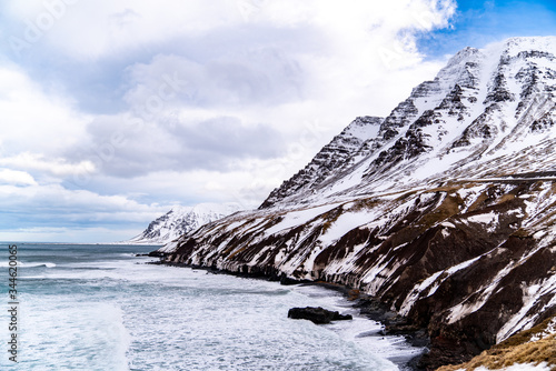 The coast of northern Iceland. A large mountain is rising from the heavy waves, and another snow covered mountain is seen in the distance.