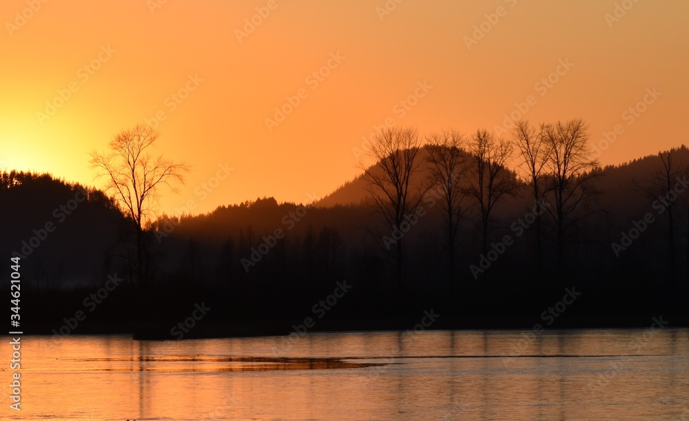 Orange and purple misty sunset, silhouette of trees in front of hills, with calm lake in foreground. Orange and purple misty sunset, silhouette of trees in front of hills, with calm lake in foreground.