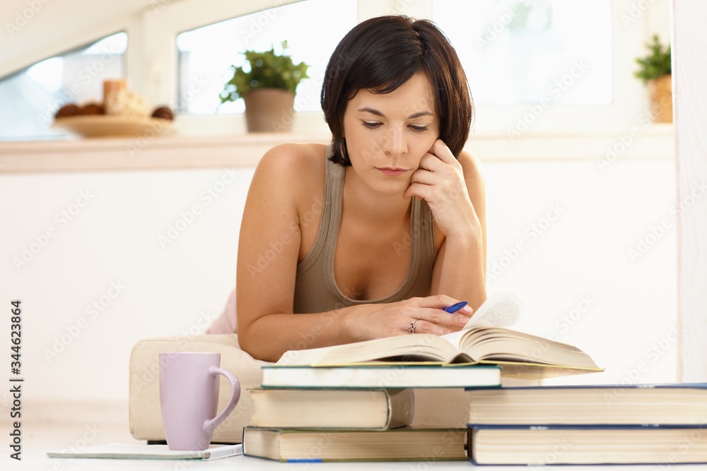 Student learning home from books. Young woman lying on floor at home in living room, writing in exercise book, reading from book, smiling. Student learning home from books. Young woman lying on floor at home in living room, writing in exercise book, reading from book, smiling.