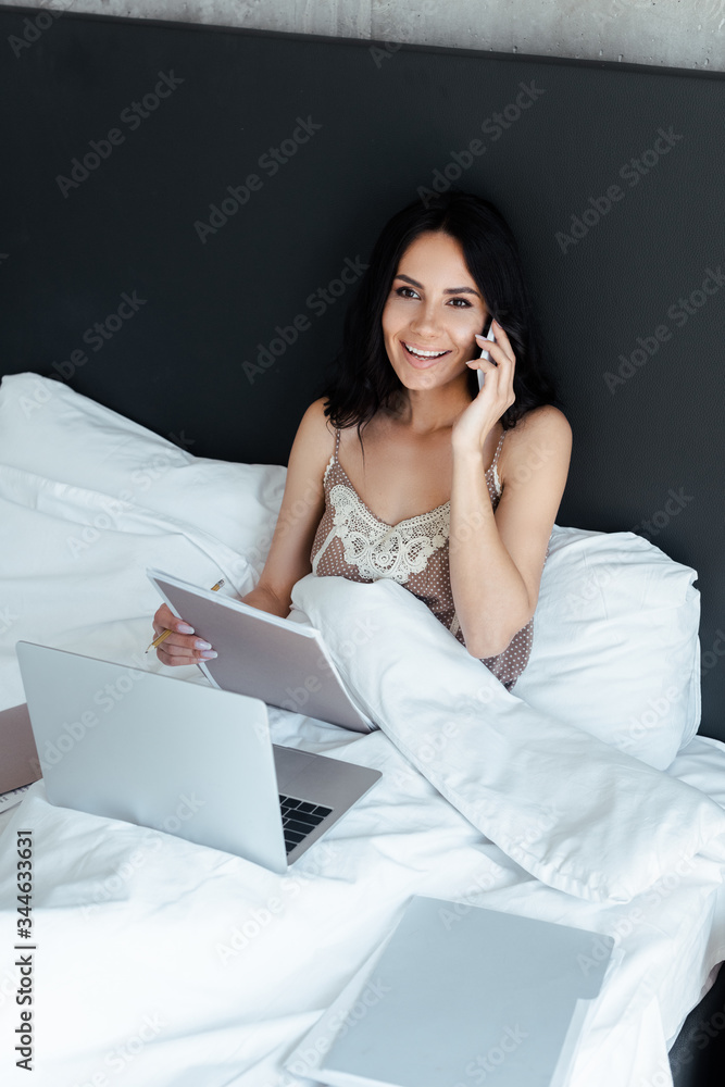 attractive woman talking on smartphone and working with business documents and laptop in bed on self isolation attractive woman talking on smartphone and working with business documents and laptop in bed on self isolation