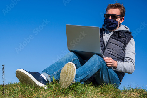 A young man in dark glasses and medical mask sits on grass in Park with laptop in his hands against blue cloudless sky. Remotely works during allergies or coronavirus pandemic. office outdoors
