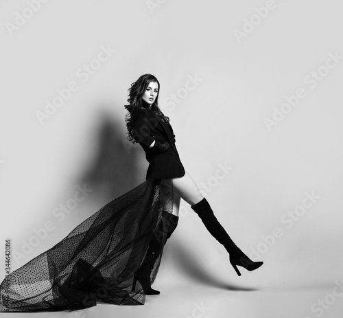 Young beautiful brunette woman in black clothes with long transparent hem and high boots making step and looking at camera