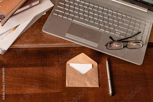 White empty postcard with craft envelope on the retro,wooden desktop with keyboard of modern laptop,black eyeglasses and grey pen.Flat lay of workplace.Copy space.Love letter to boyfreind. photo