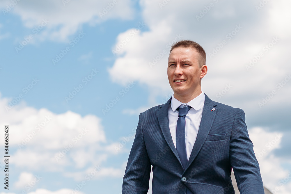 Lifestyle portrait of happy groom outdoor at nature with sky and clouds on background. Cheerful fiance with smiling face in blue jakcet