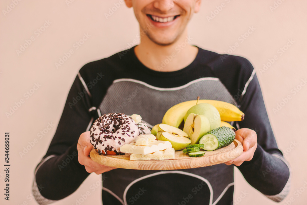 Closeup of young fitness man choosing between healthy food and unhealthy sugar snacks and smiling. Happy sports man holding wooden board with fresh fruits, vegetables, sugar donut, chocolate. Dieting Closeup of young fitness man choosing between healthy food and unhealthy sugar snacks and smiling. Happy sports man holding wooden board with fresh fruits, vegetables, sugar donut, chocolate. Dieting