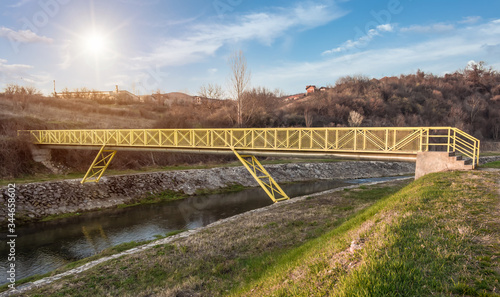 Small yellow metal pedestrian bridge outside the city. Timok river in eastern Serbia, blue sky and the sun in the background.