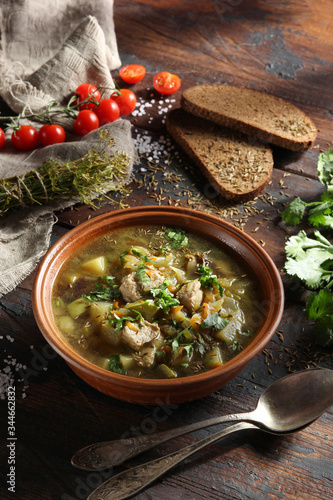 Rustic soup with vegetables, meat and fresh herbs in a clay bowl on an old wooden background. Bread, cumin, coriander, tomatoes, spoons and a bunch of dill. Background image, copy space