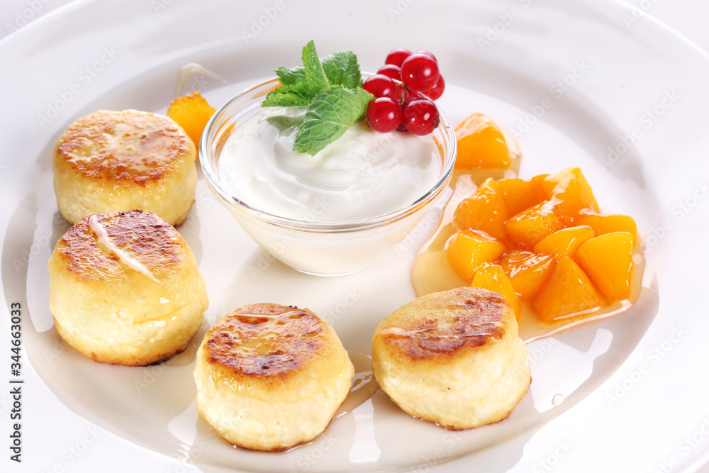Cheese cakes with sour cream, slices of sweet peach, mint and red currants on a white plate. Cheese cakes with sour cream, slices of sweet peach, mint and red currants on a white plate.
