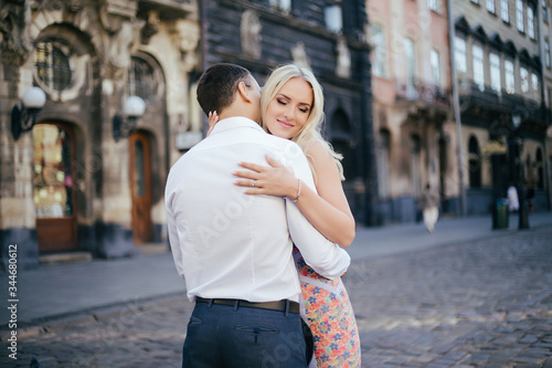 Lovely couple walking around the block. Dark-haired man in a white shirt hugging a blonde in a beautiful dress
