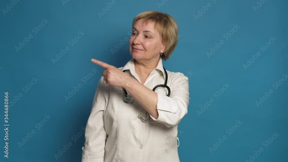 Eldery caucasian doctor lady in white coat on blue background. She wear stethoscope and pointing her fingers Eldery caucasian doctor lady in white coat on blue background. She wear stethoscope and pointing her fingers