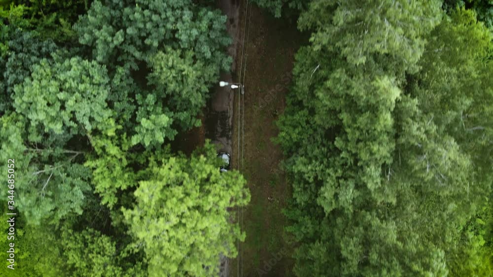 Aerial view flying over old patched two lane forest road with car moving green trees of dense woods growing both sides. Car driving along the forest road. AERIAL: Car driving through pine forest. Aerial view flying over old patched two lane forest road with car moving green trees of dense woods growing both sides. Car driving along the forest road. AERIAL: Car driving through pine forest.