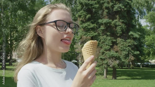 Cute blonde girl in glasses licking palatable ice cream in a waffle cone with delight on hot summer day looking away from camera.