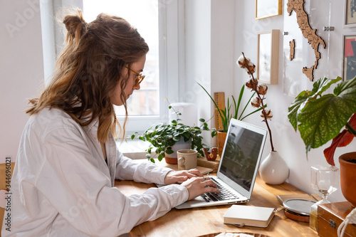 Young woman freelancer/designer working on computer from home office during self-isolation due to coronavirus. Cozy workplace surrounded by plants. Remote work, Telecommuting, Distance job. 