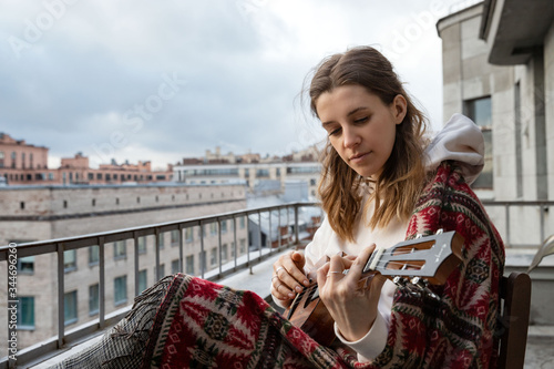 Young caucasian hipster/hippie woman in casual clothes playing Hawaiian guitar, sings a song on a ukulele during self-isolation in the house on terrace. Hobby. Daily life in your own home.  © DimaBerlin