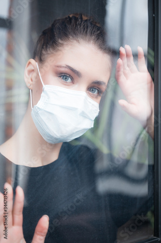 An Asian woman in a medical mask with her palms rests on the glass and looks sadly out the window. Quarantine and home isolation.