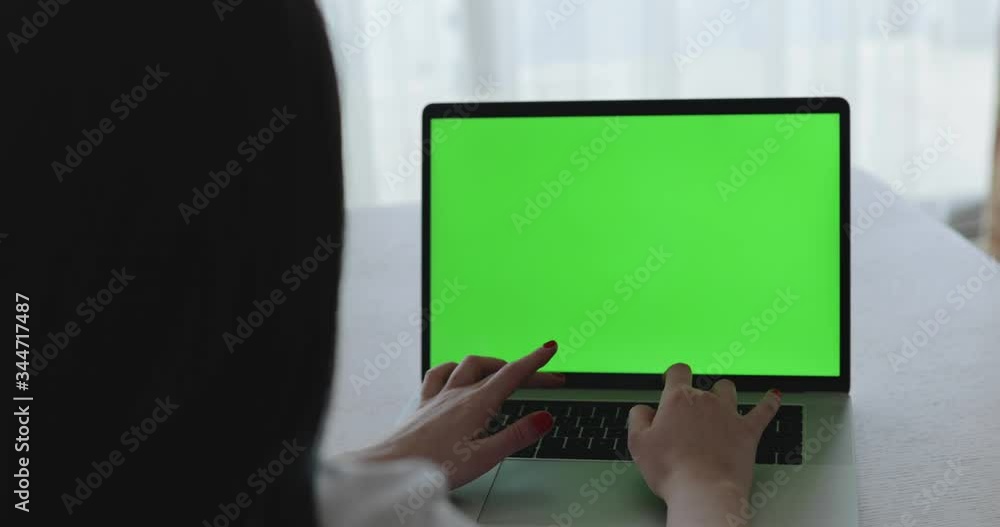 Female person typing on laptop keyboard with green screen on table at window Female person typing on laptop keyboard with green screen on table at window